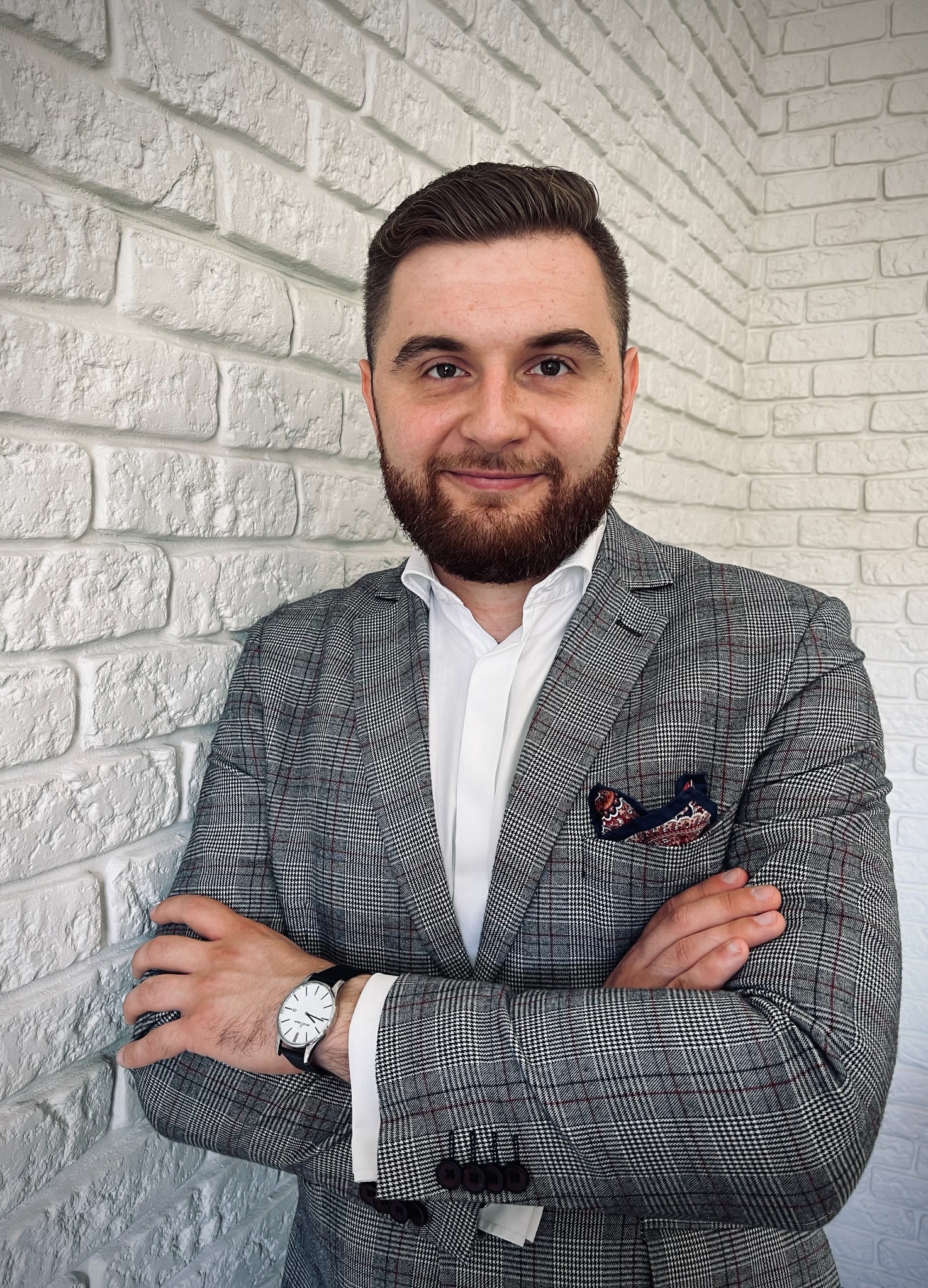 Portret mężczyzny z brodą w szarej marynarce w kratę, stojącego na tle białej ściany z cegieł. Na nadgarstku widoczny zegarek. W kieszonce marynarki ozdobna poszetka.