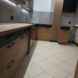 Nowoczesna kuchnia z drewnianymi frontami szafek dolnych i grafitowymi górnymi, szarymi płytkami na ścianie, drewnianymi lamelami dekoracyjnymi i beżową podłogą z płytek.