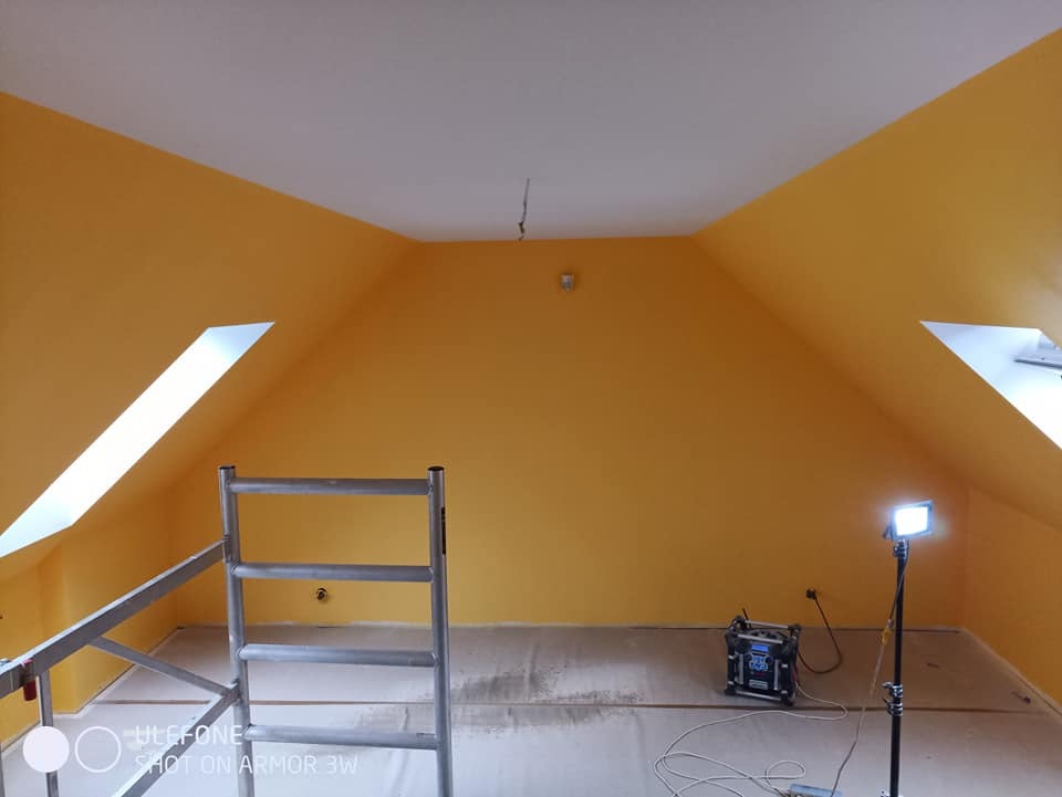 Pomalowane na żółto poddasze ze skosami i oknami dachowymi, widoczny metalowy ruszt, lampa oświetleniowa i agregat malarski.