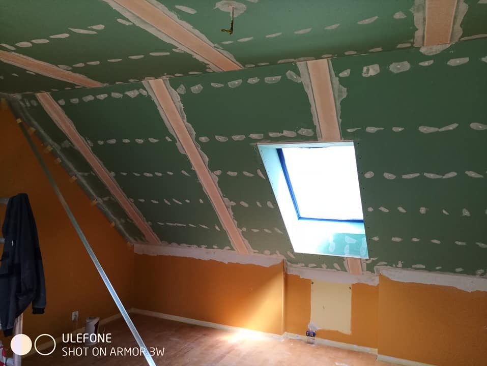 Poddasze w trakcie prac wykończeniowych: zielone płyty gipsowo-kartonowe zamocowane na skosach, widoczne łączenia szpachlowane, okno dachowe w ramie, pomarańczowe ściany w dolnej części, widoczny...