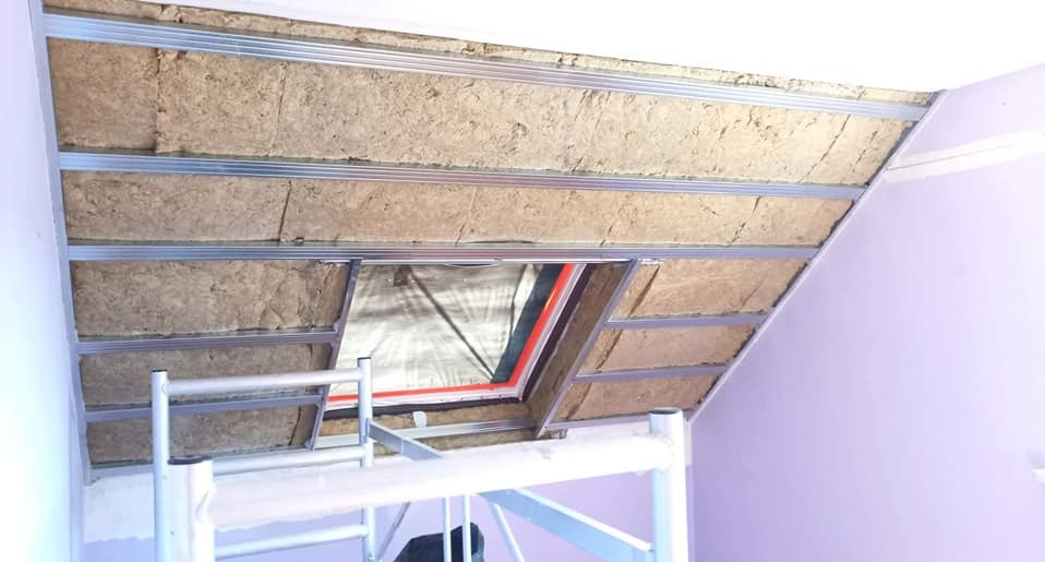 Skośny sufit poddasza z widoczną izolacją z wełny mineralnej, zamontowaną między metalowymi profilami, z oknem dachowym w trakcie montażu, widok ze stojącego rusztowania.