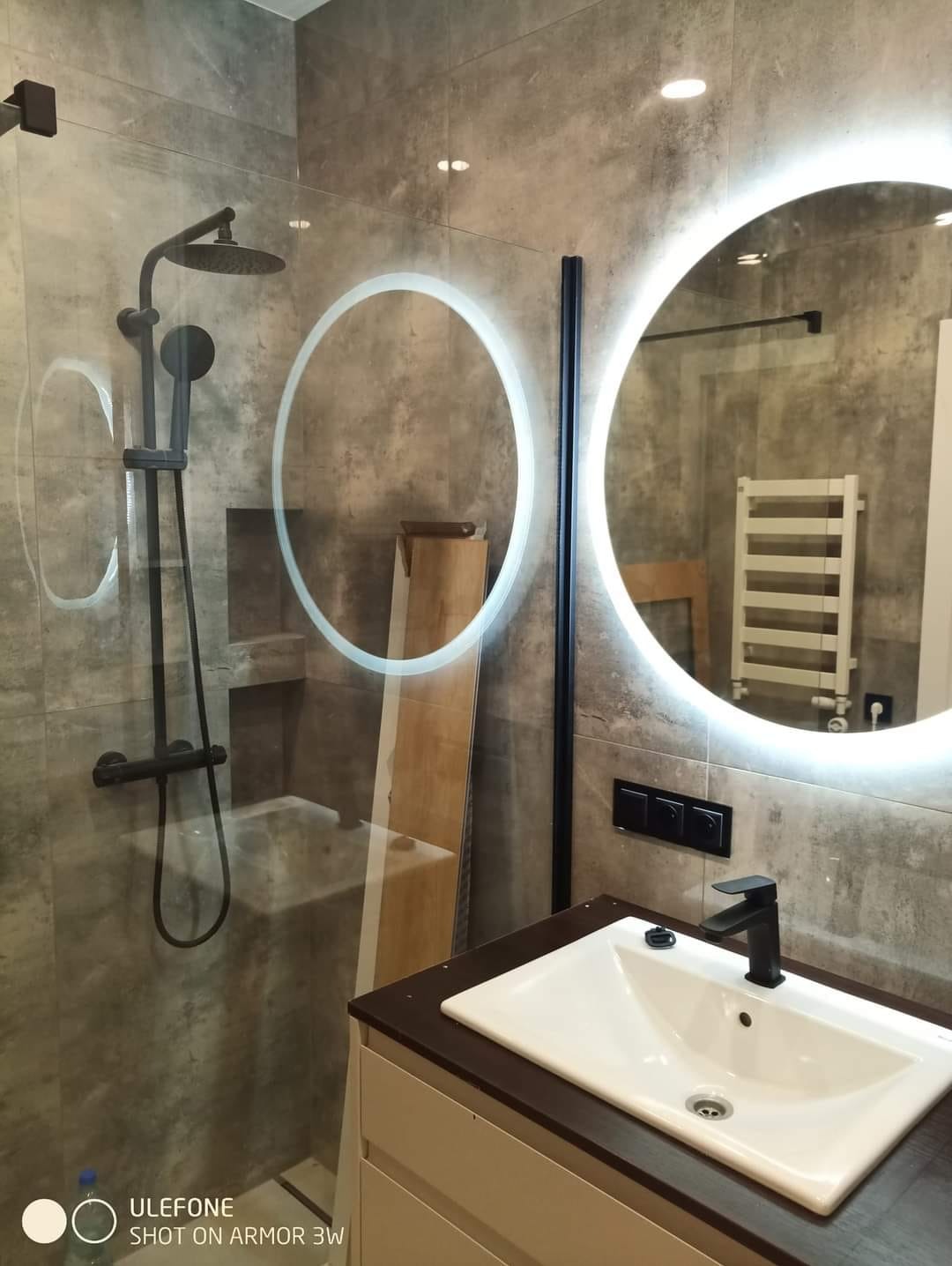 Nowoczesna łazienka z szarą płytką imitującą beton, okrągłe lustro z podświetleniem LED, czarna armatura i kabina prysznicowa typu walk-in z widoczną instalacją hydrauliczną w kolorze czarnym.