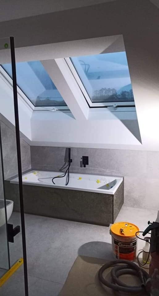 Nowoczesna łazienka na poddaszu z wanną obudowaną płytkami imitującymi kamień, czarną armaturą i oknem dachowym, widok na krajobraz za oknem.