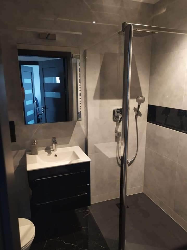 Nowoczesna łazienka po remoncie: kabina prysznicowa ze szklaną ścianką, umywalka z szafką w kolorze czarnym, grafitowa podłoga z marmurowym wzorem i szare płytki na ścianach.