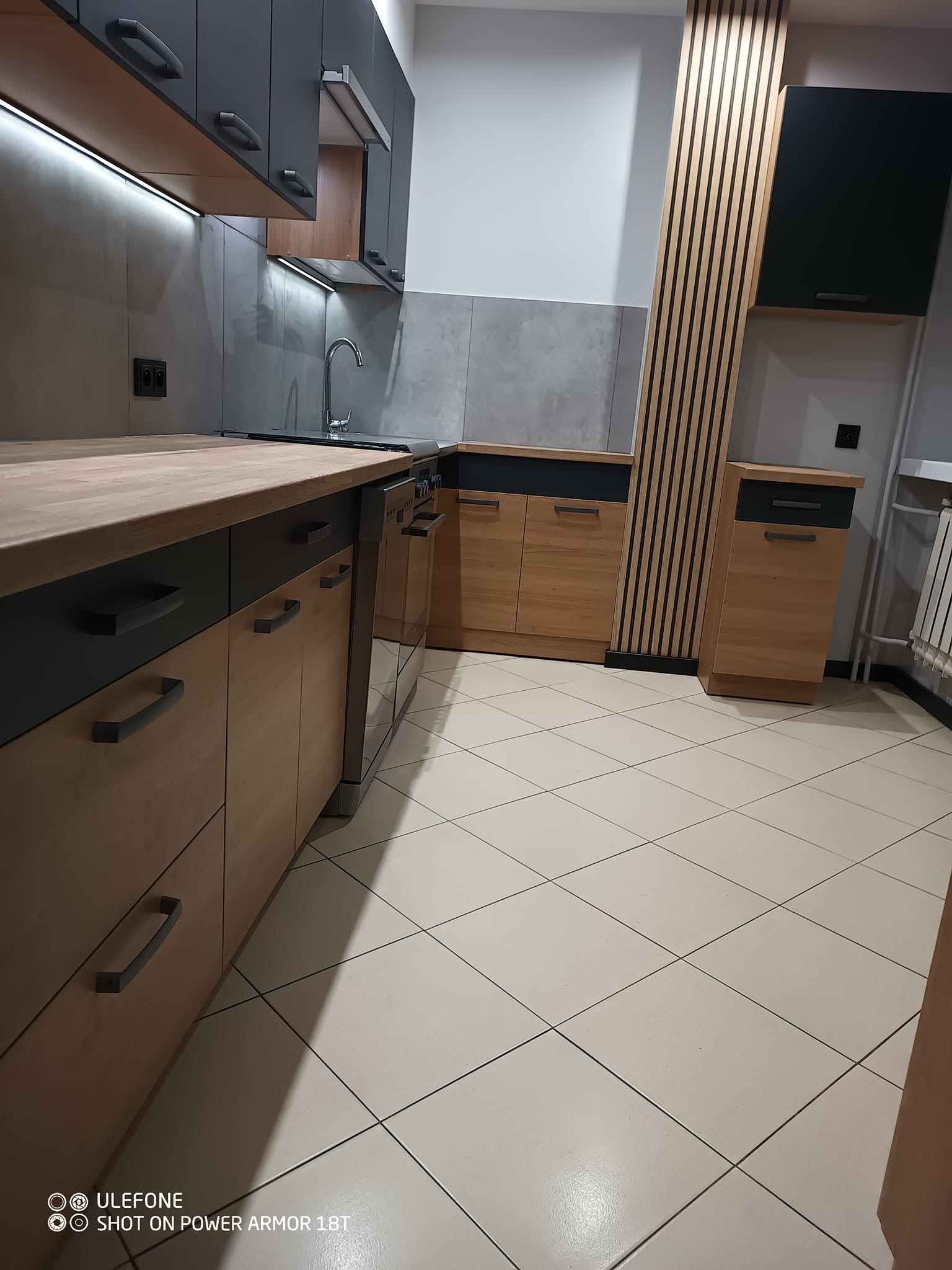 Nowoczesna kuchnia z drewnianymi frontami szafek dolnych i grafitowymi górnymi, szarymi płytkami na ścianie, drewnianymi lamelami dekoracyjnymi i beżową podłogą z płytek.