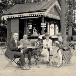 Czarno-białe zdjęcie z lat 20-tych przedstawiające cztery osoby siedzące przy stolikach przed budką z napojami 'Mousse Dilmant' w parku, mężczyzna w garniturze trzyma kieliszek, kelnerka w tle.