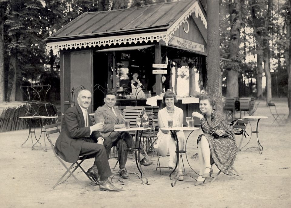 Czarno-białe zdjęcie z lat 20-tych przedstawiające cztery osoby siedzące przy stolikach przed budką z napojami 'Mousse Dilmant' w parku, mężczyzna w garniturze trzyma kieliszek, kelnerka w tle.