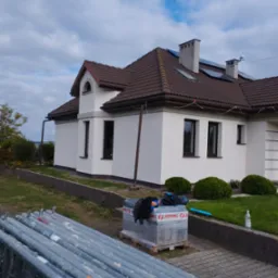 Nowo otynkowany, biały dom z brązowym dachem, widoczne rusztowania i materiały izolacyjne na palecie, przygotowane do dalszych prac.