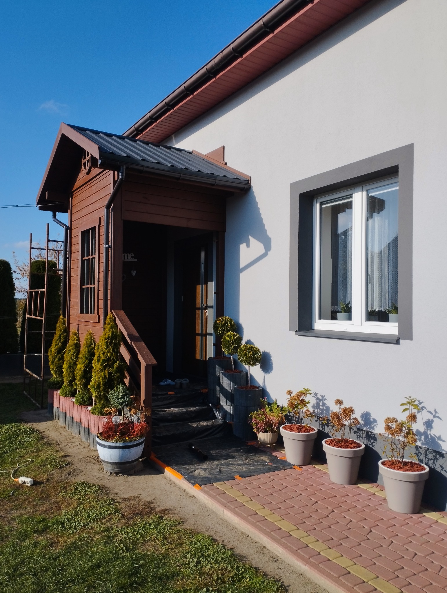 Jasnoszary dom z drewnianą werandą i szarą ramą okienną, widoczne donice z roślinami, zabezpieczona folią ścieżka przed wejściem.