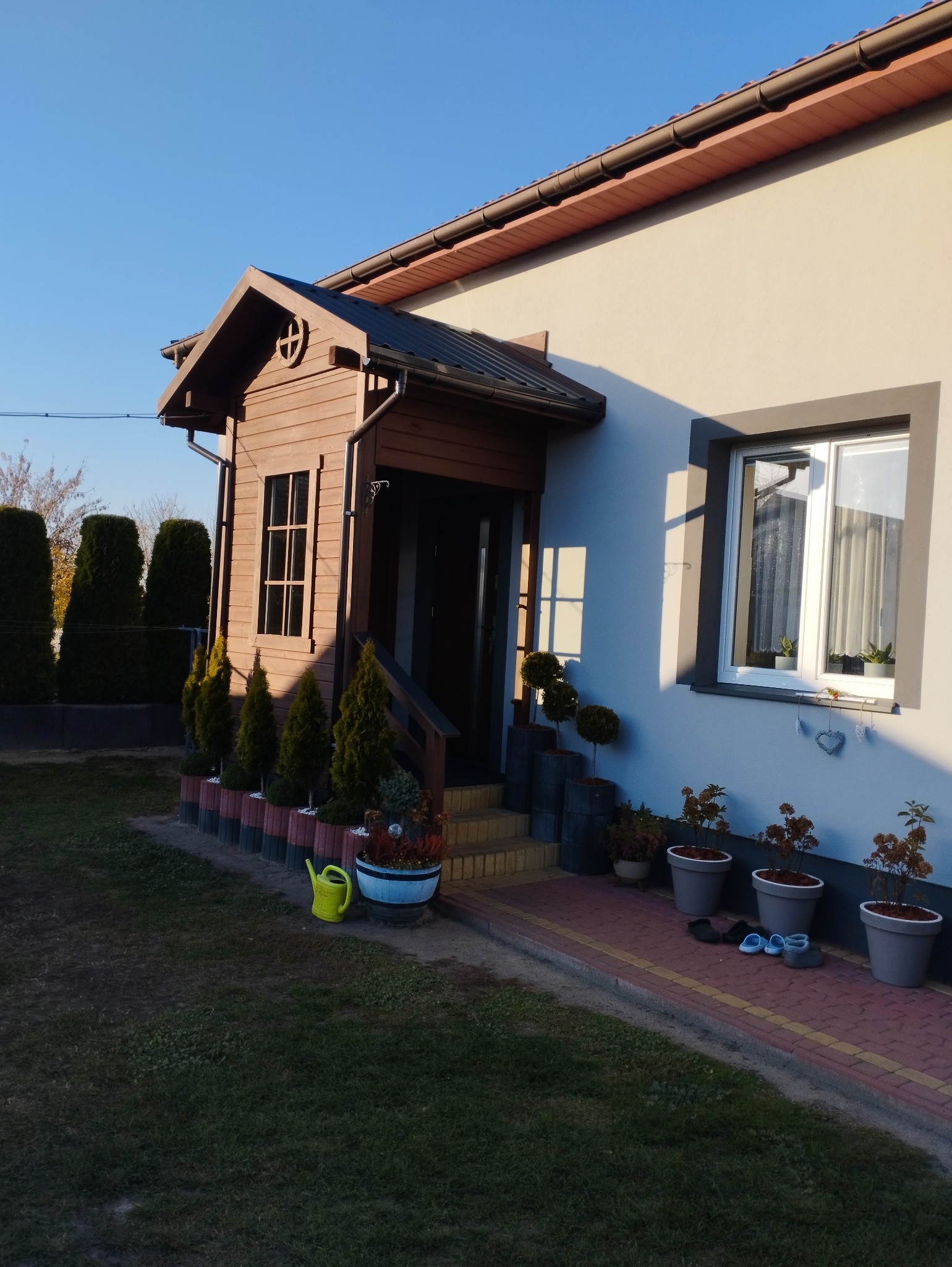 Ocieplony dom jednorodzinny z drewnianą werandą i świeżo pomalowaną elewacją w odcieniach beżu i szarości, z dekoracyjnymi roślinami w donicach i żółtą konewką w ogrodzie.