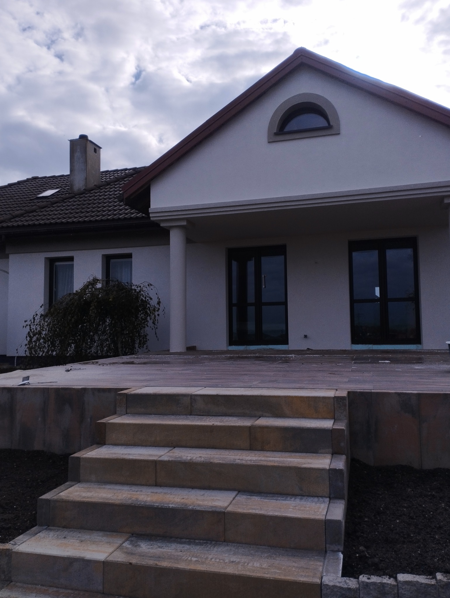 Nowoczesny dom jednorodzinny z ocieploną elewacją w kolorze jasnego beżu, widoczne dwuskrzydłowe okna z czarnymi ramami, kamienne schody prowadzące na taras i fragment ogrodu z młodym drzewkiem.