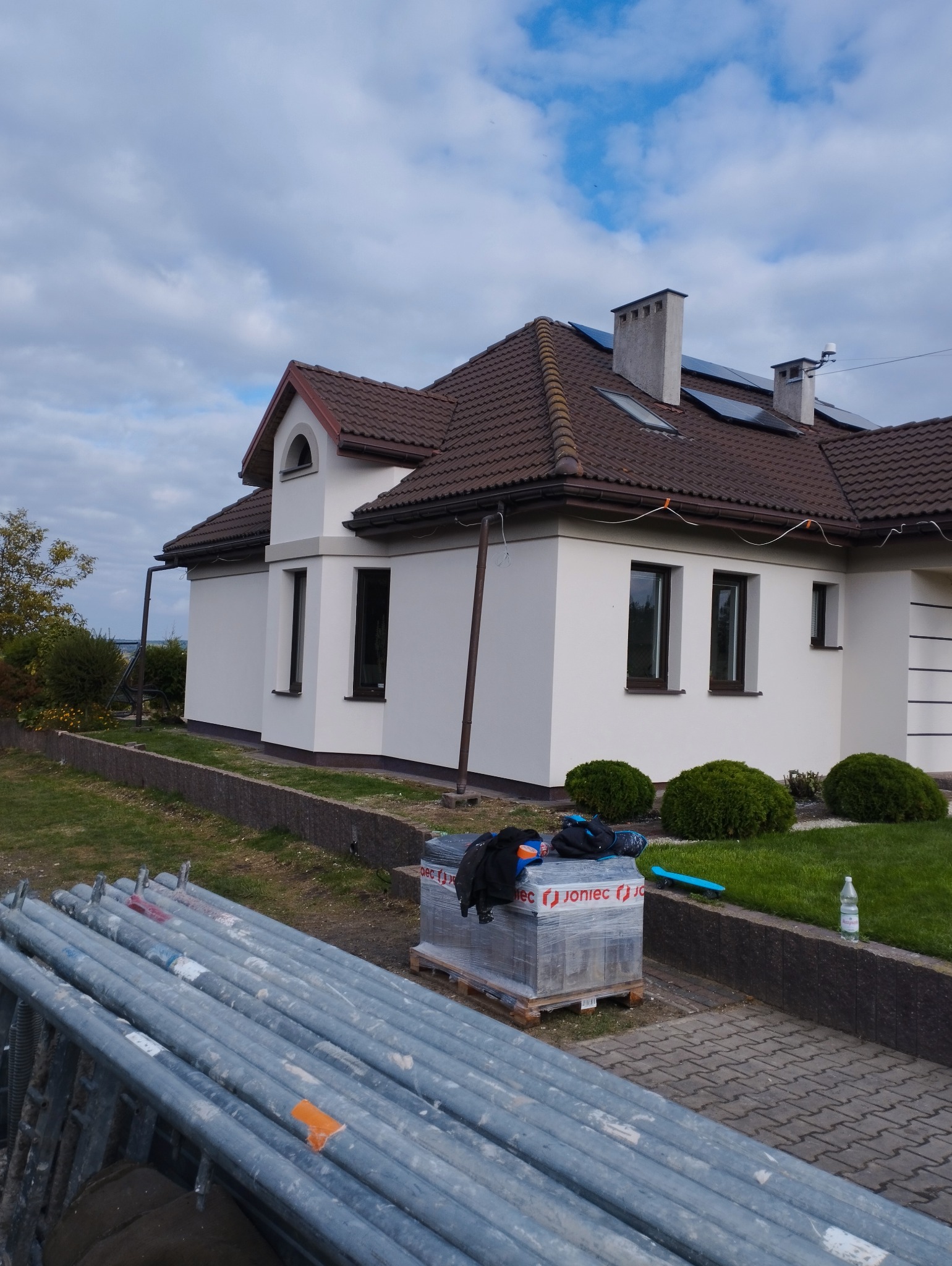 Nowo otynkowany, biały dom z brązowym dachem, widoczne rusztowania i materiały izolacyjne na palecie, przygotowane do dalszych prac.