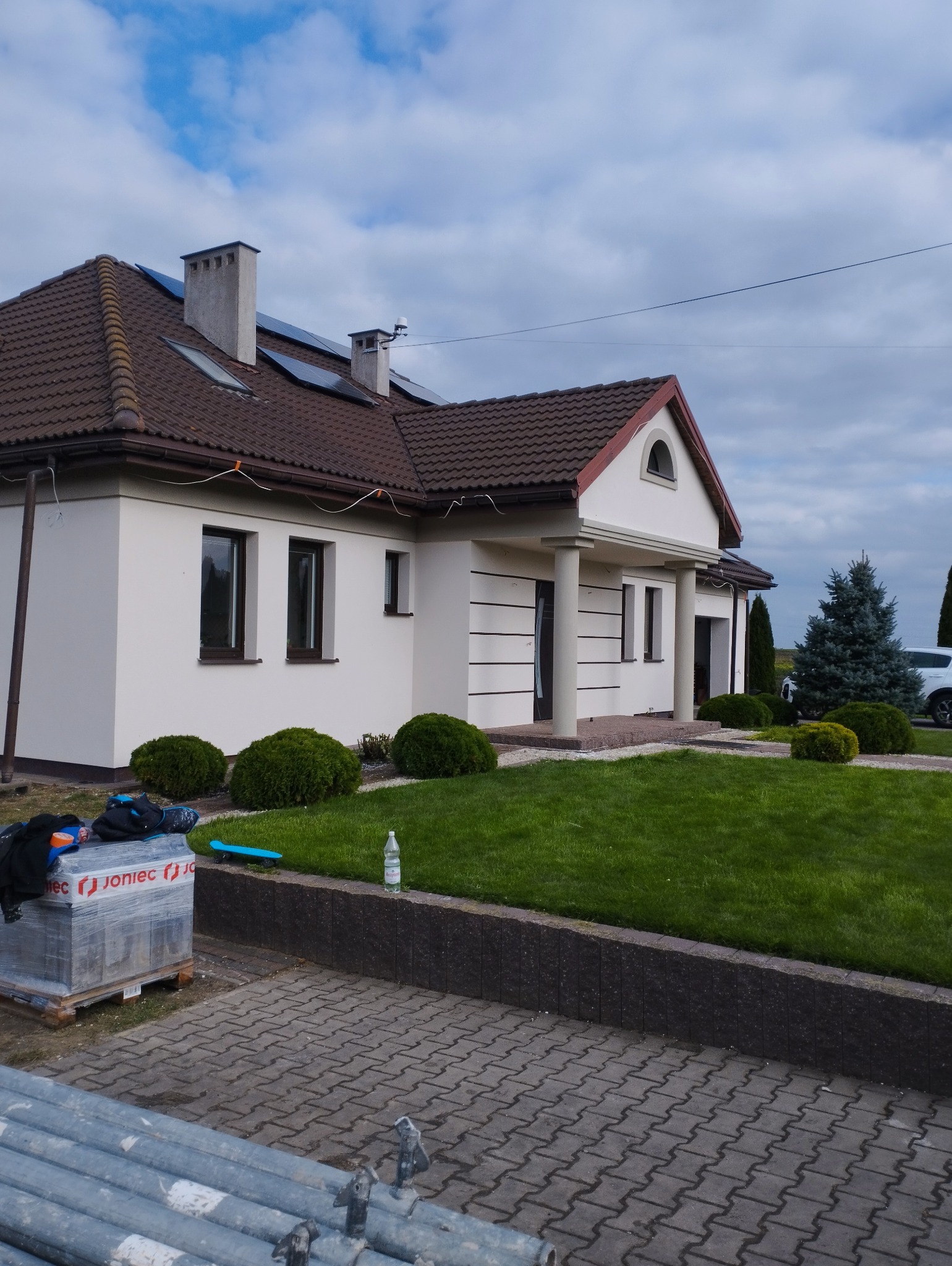 Jasny, ocieplony dom jednorodzinny z brązowym dachem i panelami słonecznymi, widok z zewnątrz na trawnik, dekoracyjne krzewy i rusztowanie w tle.