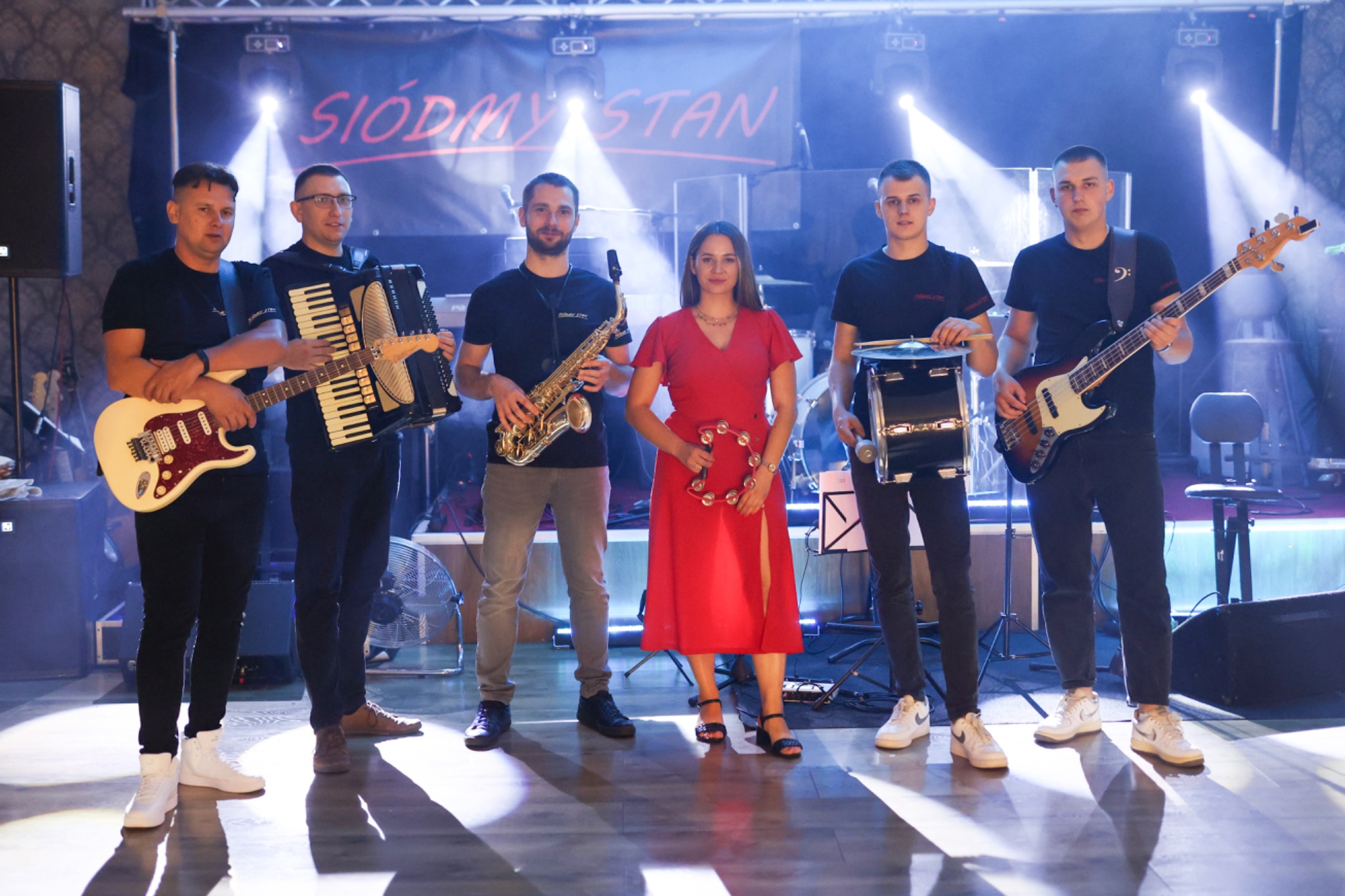 Zespół muzyczny w składzie sześcioosobowym, z gitarą elektryczną, akordeonem, saksofonem, tamburynem, perkusją i gitarą basową, na scenie z oświetleniem i banerem z nazwą zespołu.