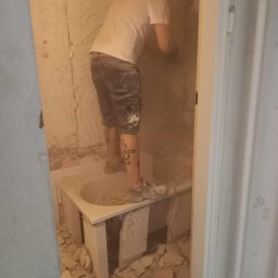 AleksanderBorsukUsługiRemontowe - Mężczyzna w krótkich spodenkach z tatuażem na łydce stoi w wannie, skuwa starą glazurę ze ściany w zapylonej łazience podczas remontu.