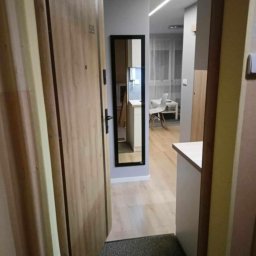 AleksanderBorsukUsługiRemontowe - Otwarta, drewniana drzwi wejściowe z numerem 59, widok na przedpokój z lustrem w czarnej ramie, podłoga wyłożona panelami, w tle widoczny fragment kuchni z białymi meblami i krzesłem.