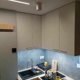 AleksanderBorsukUsługiRemontowe - Wykończona kuchnia w Bochni: szare szafki wiszące z podświetleniem LED nad blatem z czarnym zlewem i płytą indukcyjną, widoczny fragment lodówki.