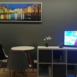 AleksanderBorsukUsługiRemontowe - Pokój z szarą ścianą, na której wisi obraz z mostem Rialto w Wenecji, biały zegar oraz włączony telewizor na białej szafce, obok stoi mały biały stolik z dwoma krzesłami.