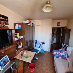 AleksanderBorsukUsługiRemontowe - Zdjęcie wnętrza pokoju przed remontem, widoczne stare meble, brudne ściany i podłoga z paneli w kwadraty, przez otwarte drzwi widać osobę w innym pomieszczeniu.