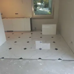 Białe, marmurowe płytki podłogowe z czarnymi akcentami ułożone w pomieszczeniu w trakcie prac wykończeniowych; widoczne niedokończone ściany, białe szafki kuchenne bez blatów i okno.