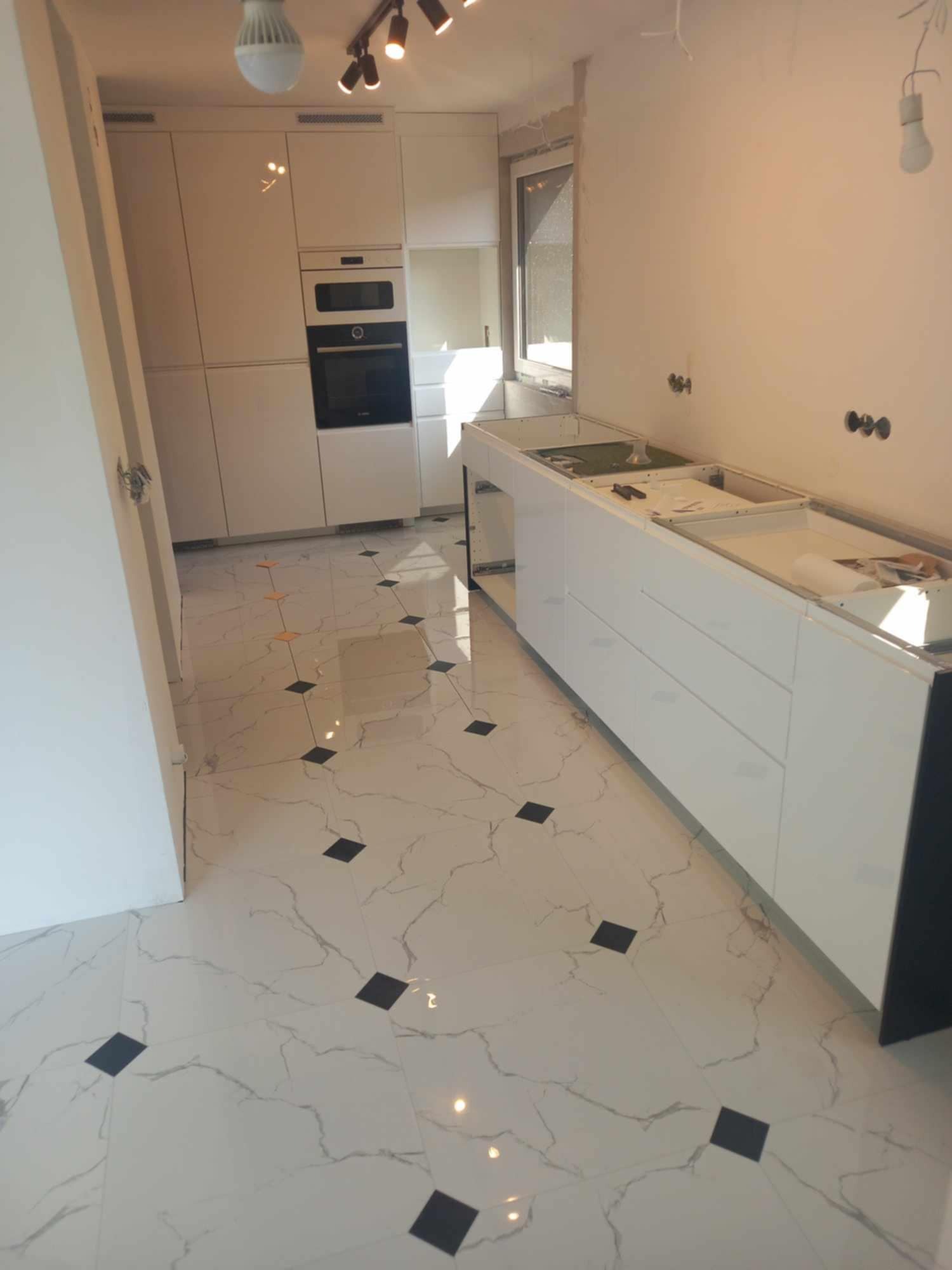 Wnętrze kuchni w trakcie montażu: białe szafki kuchenne bez blatów, marmurowe płytki podłogowe z czarnymi akcentami, widoczne przygotowania do instalacji oświetlenia.