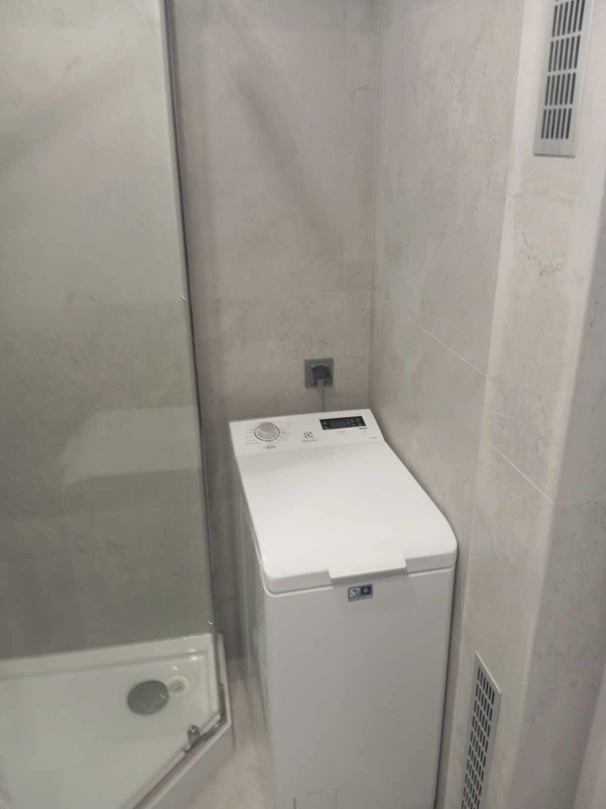 Kompaktowa łazienka z kabiną prysznicową i pralką ładowaną od góry, wykończona jasnymi, imitującymi kamień płytkami ceramicznymi z subtelną fakturą.