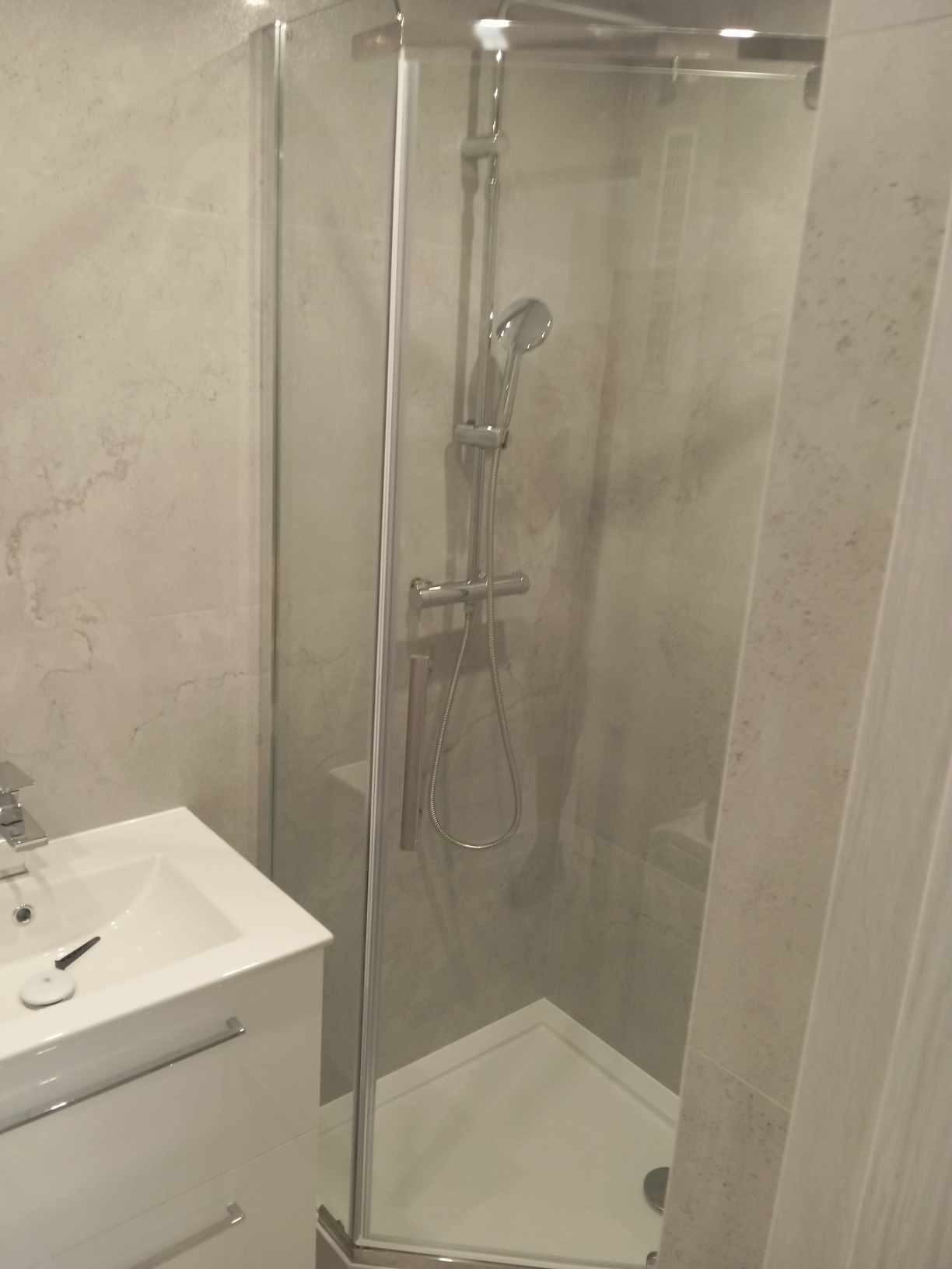 Nowoczesna kabina prysznicowa z deszczownicą i baterią termostatyczną, narożna, ze szklanymi drzwiami, obok biała umywalka z szafką, szare płytki imitujące marmur na ścianach.