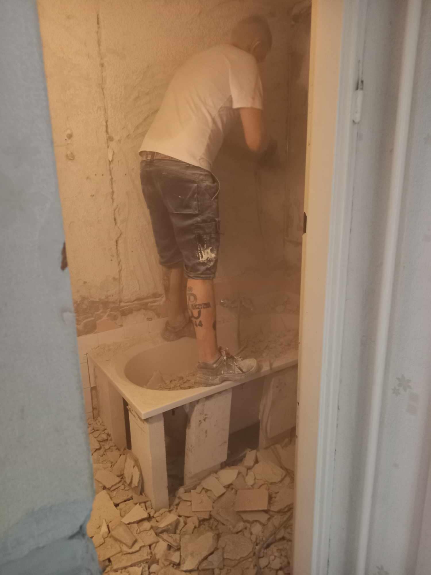 Mężczyzna w krótkich spodenkach z tatuażem na łydce stoi w wannie, skuwa starą glazurę ze ściany w zapylonej łazience podczas remontu.