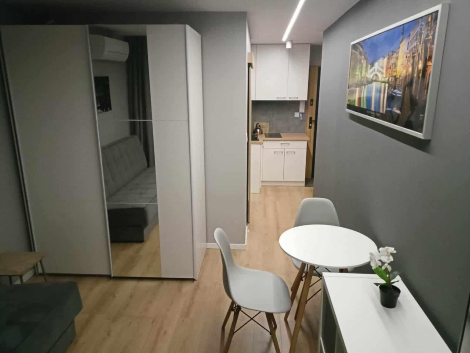 Wnętrze małego mieszkania z aneksem kuchennym, szafą z lustrzanymi drzwiami, okrągłym stołem i dwoma krzesłami oraz obrazem na szarej ścianie.