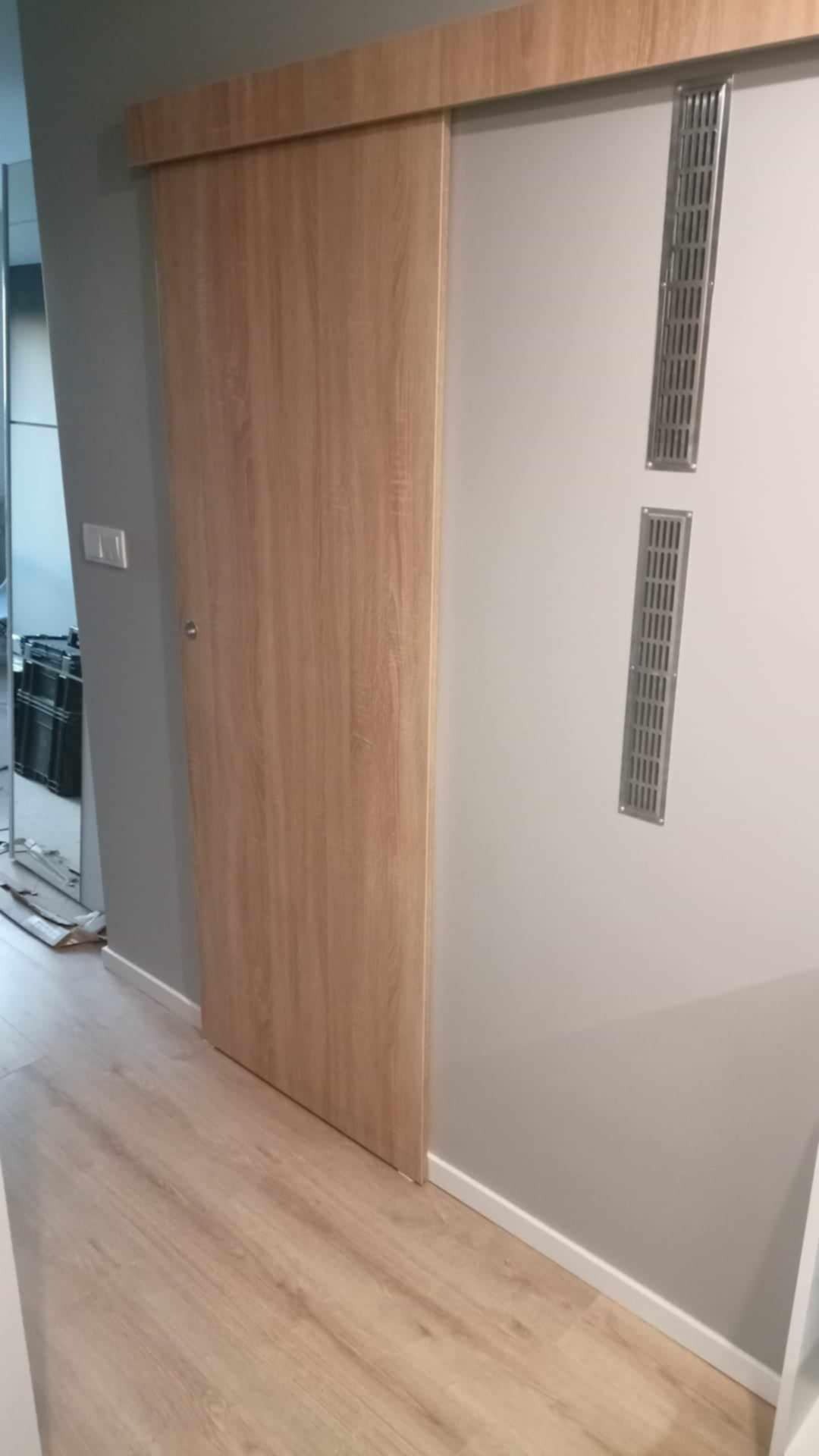 Zamknięte drewniane drzwi przesuwne z widocznymi kratkami wentylacyjnymi na szarej ścianie i jasnymi panelami podłogowymi.