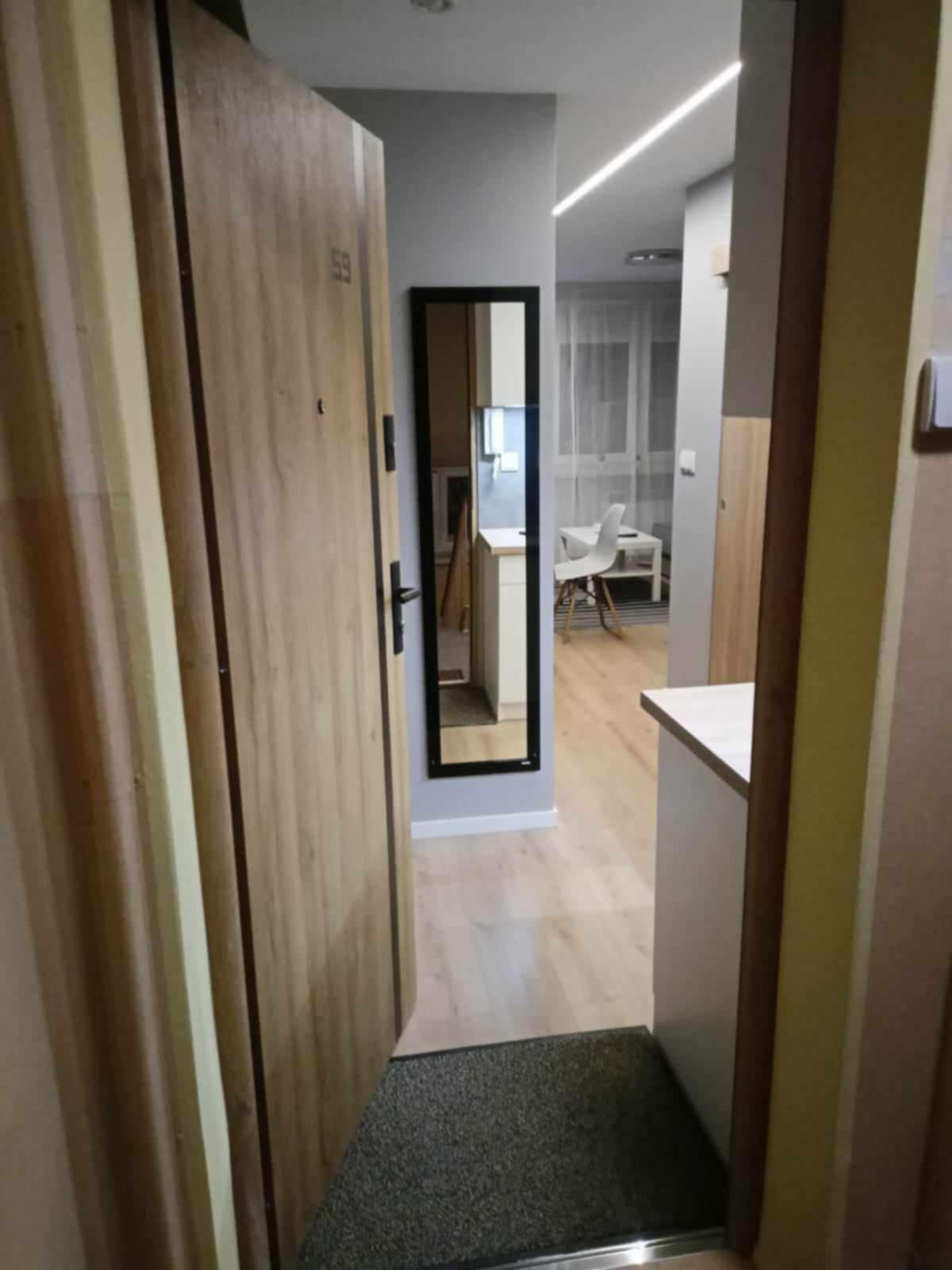 Otwarta, drewniana drzwi wejściowe z numerem 59, widok na przedpokój z lustrem w czarnej ramie, podłoga wyłożona panelami, w tle widoczny fragment kuchni z białymi meblami i krzesłem.
