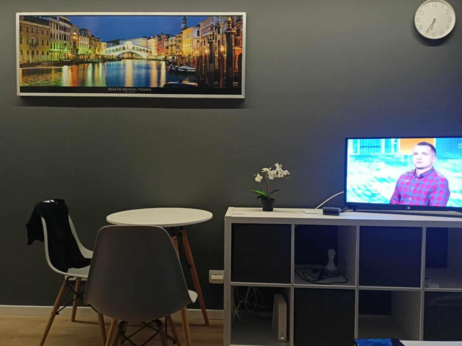 Pokój z szarą ścianą, na której wisi obraz z mostem Rialto w Wenecji, biały zegar oraz włączony telewizor na białej szafce, obok stoi mały biały stolik z dwoma krzesłami.