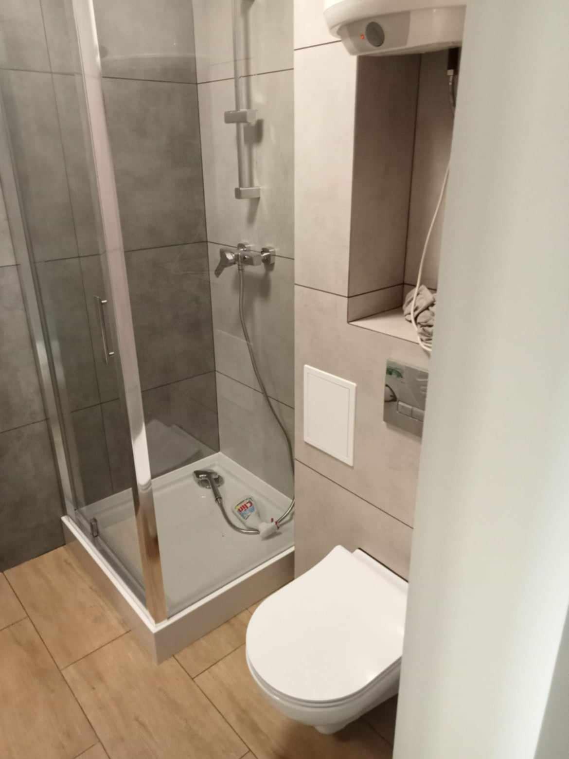 Szara łazienka z kabiną prysznicową, podwieszaną toaletą i drewnopodobną podłogą; w kabinie widoczny środek czyszczący.