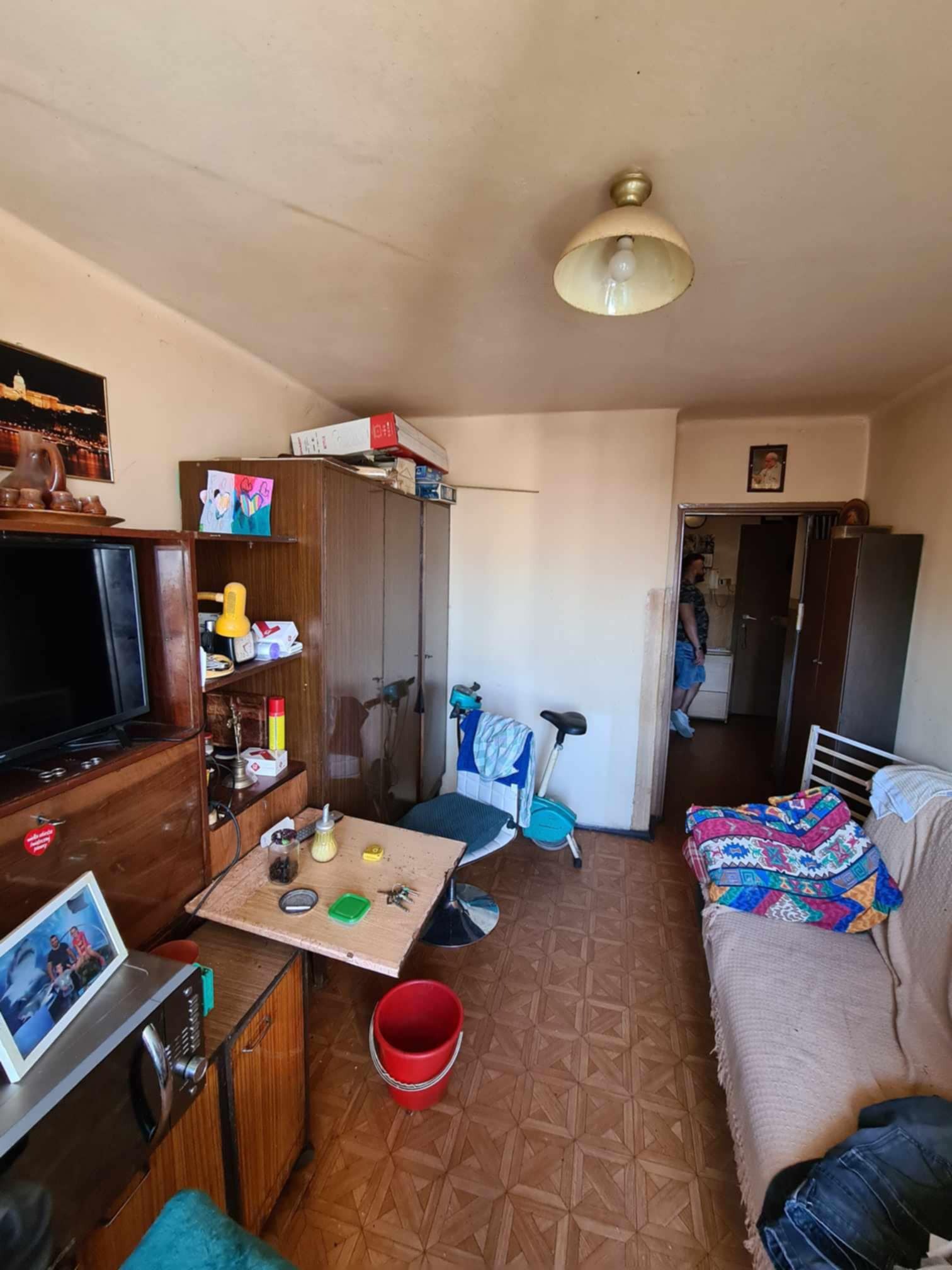 Zdjęcie wnętrza pokoju przed remontem, widoczne stare meble, brudne ściany i podłoga z paneli w kwadraty, przez otwarte drzwi widać osobę w innym pomieszczeniu.