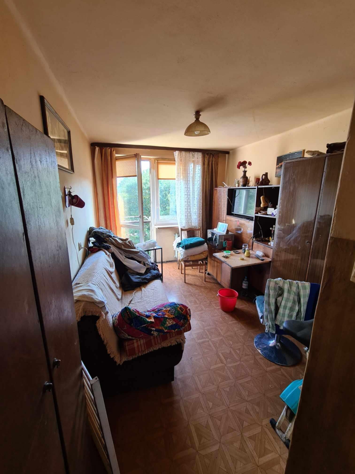 Pokój w bloku z meblami z lat 80-tych, widoczne stare szafy, kanapa z narzutą, telewizor na półce, otwarte okno z balkonem i podłoga parkietowa z wzorem.