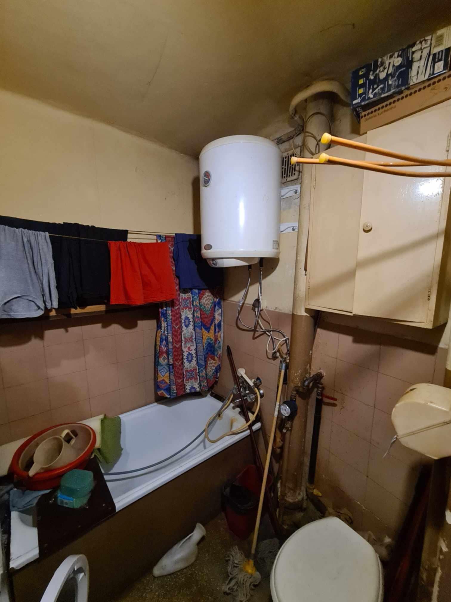 Niefotogeniczna łazienka z wiszącym bojlerem, praniem na sznurku, wanną i widocznymi rurami oraz instalacjami; widok na wnętrze przed remontem.