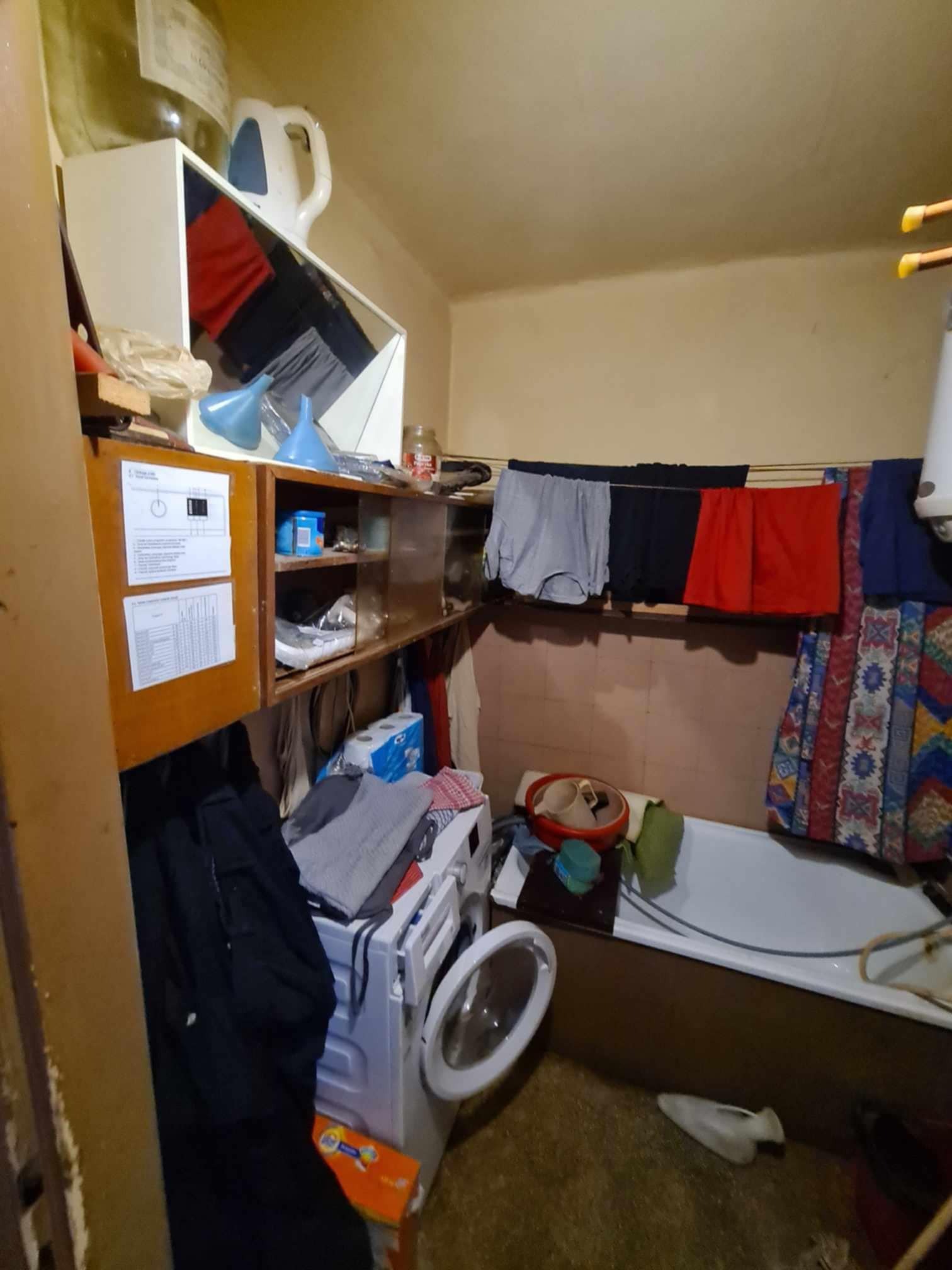 Mała, zagracona łazienka z pralką, wanną i suszącym się praniem na sznurku. Na ścianach widoczne półki z różnymi przedmiotami, w tym butelki, pudełka i akcesoria łazienkowe. Podłoga brudna,...