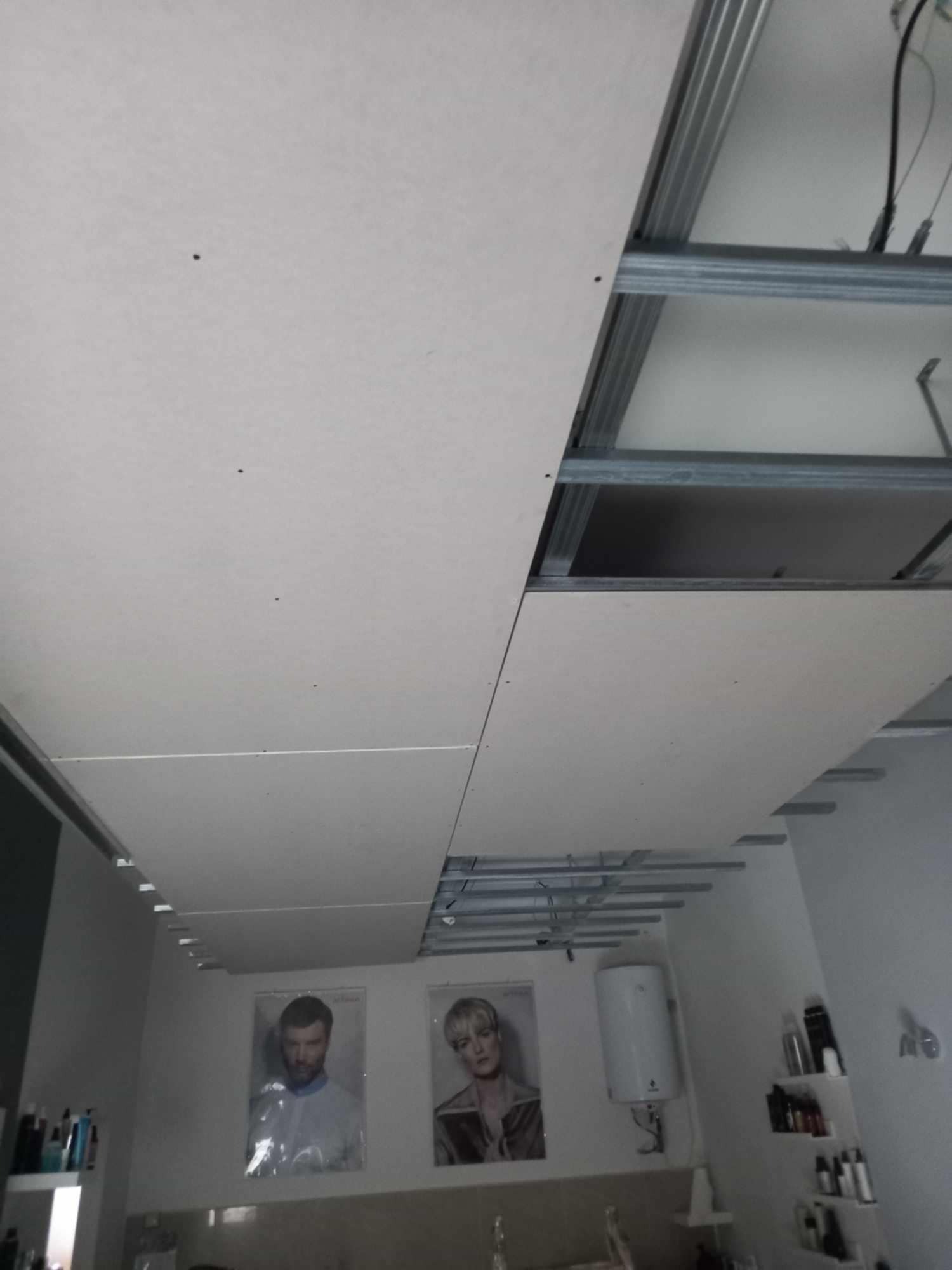 Montaż sufitu podwieszanego z płyt kartonowo-gipsowych na metalowej konstrukcji w pomieszczeniu z elementami wyposażenia salonu fryzjerskiego w tle.