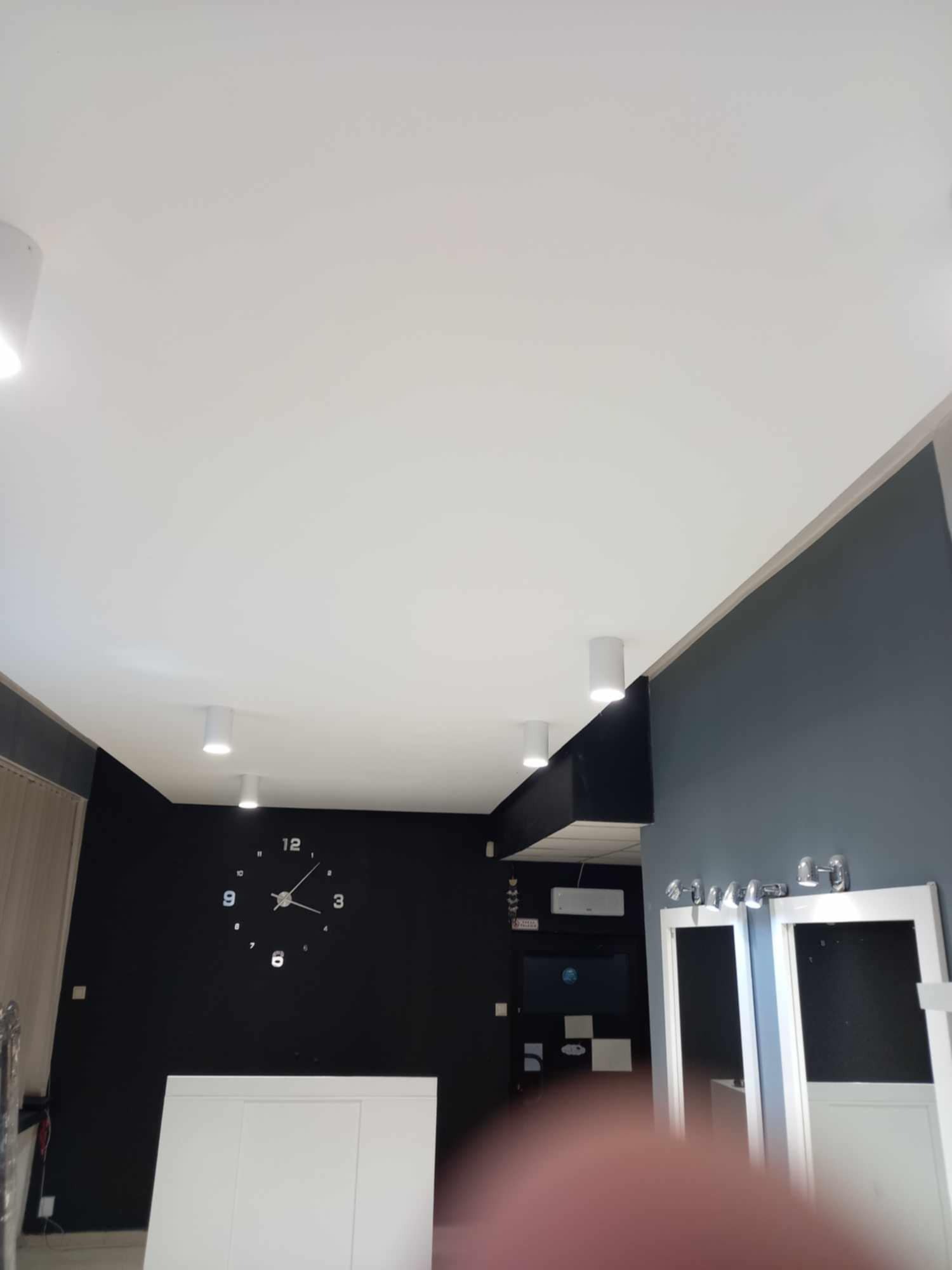 Nowoczesne wnętrze z białym sufitem podwieszanym, oświetleniem punktowym, ciemną ścianą z zegarem i elementami zabudowy w kolorze białym.