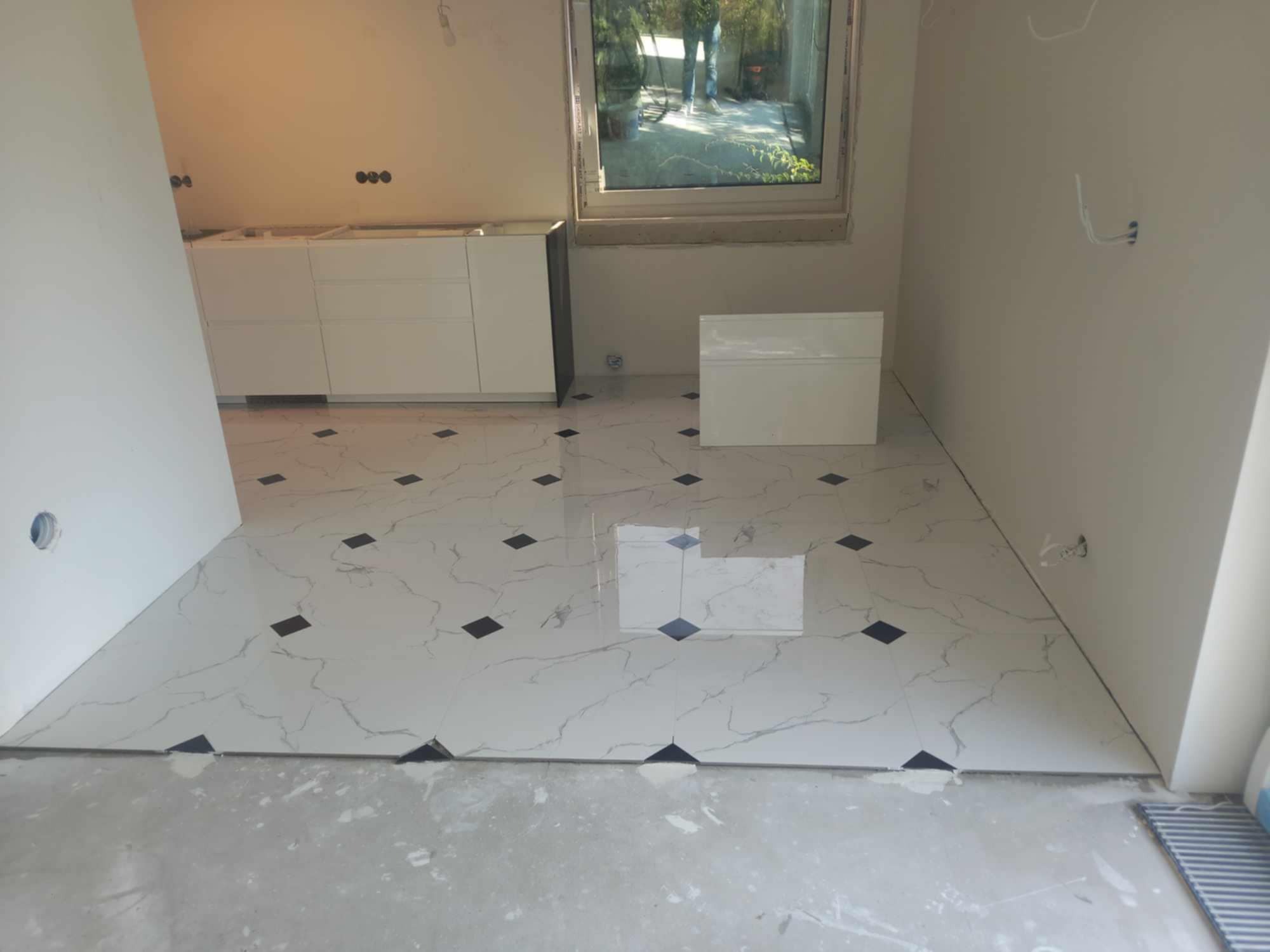 Białe, marmurowe płytki podłogowe z czarnymi akcentami ułożone w pomieszczeniu w trakcie prac wykończeniowych; widoczne niedokończone ściany, białe szafki kuchenne bez blatów i okno.