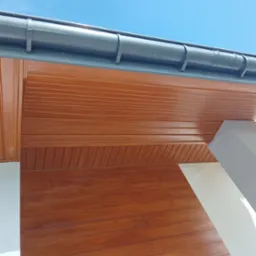 Brązowa boazeria imitująca drewno na podbitce dachu i elewacji, widoczny fragment rynny i błękitne niebo.