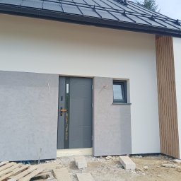 Usługi remontowo-budowlane Paweł Chruszcz - Nowoczesna elewacja domu jednorodzinnego z szarą strukturą tynku wokół drzwi wejściowych i małego okna, uzupełniona drewnianymi panelami pionowymi po prawej stronie, widoczne materiały budowlane...