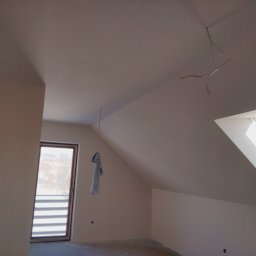 Usługi remontowo-budowlane Paweł Chruszcz - Poddasze po szpachlowaniu i malowaniu na biało, widoczne okno dachowe i wiszący ręcznik. Wystające kable elektryczne. Świeżo wykończone ściany i sufit.