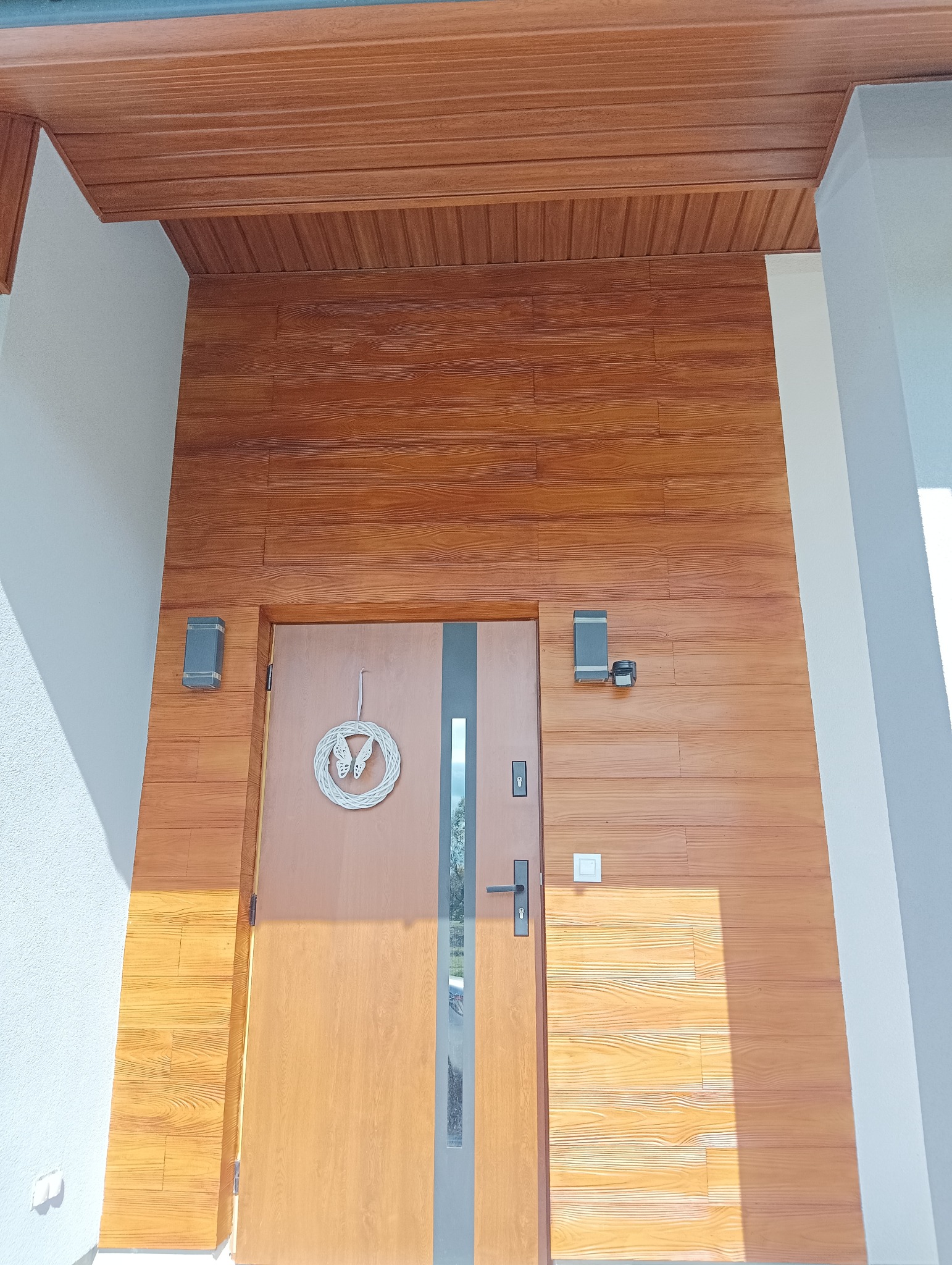 Wejście do domu z drewnopodobną elewacją, oświetleniem zewnętrznym i ozdobnym wiankiem na drzwiach.