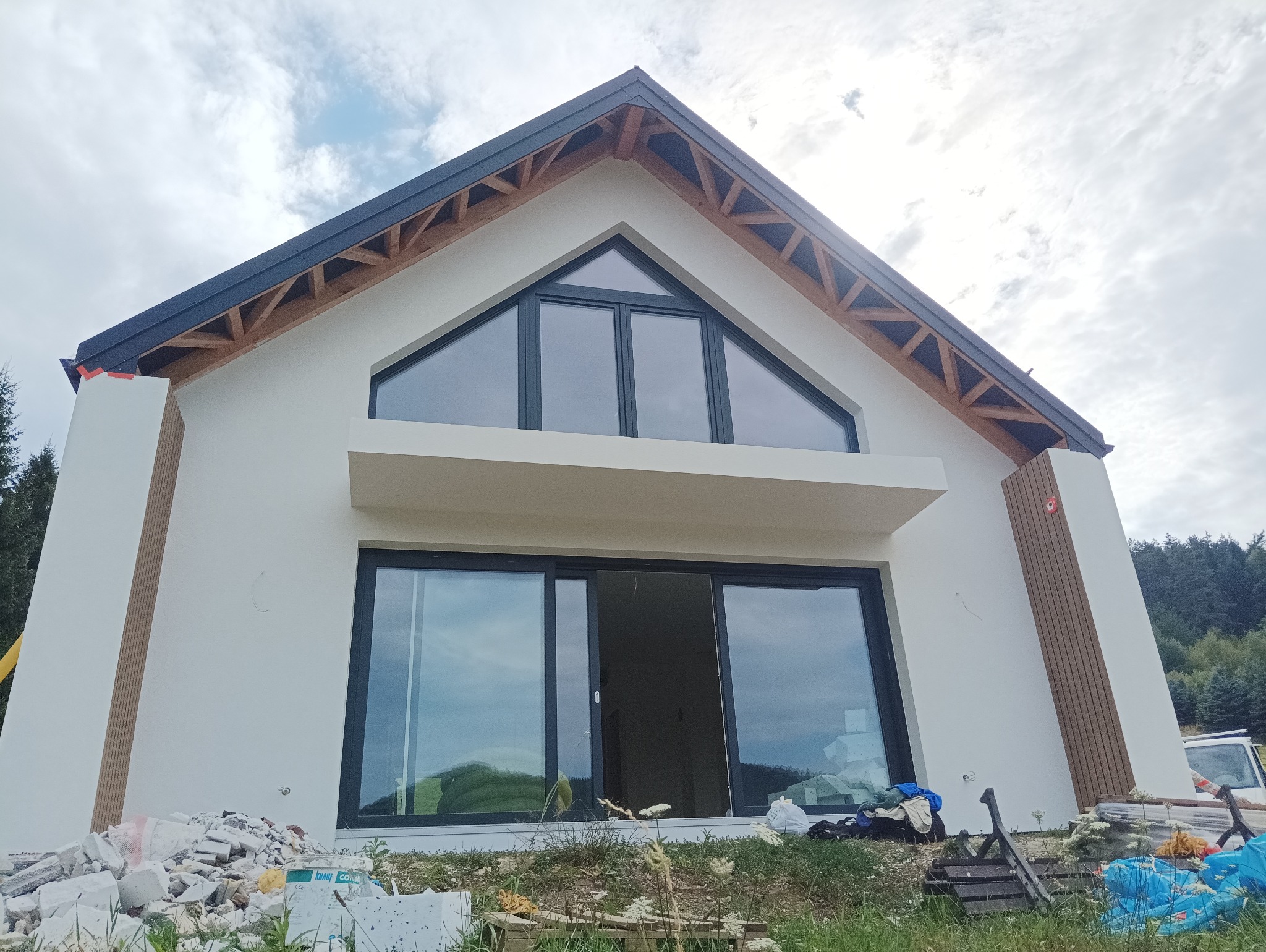 Nowoczesny dom z białymi ścianami, dużymi oknami z czarnymi ramami i drewnianymi elementami elewacji, widoczne ślady prac budowlanych na trawniku.