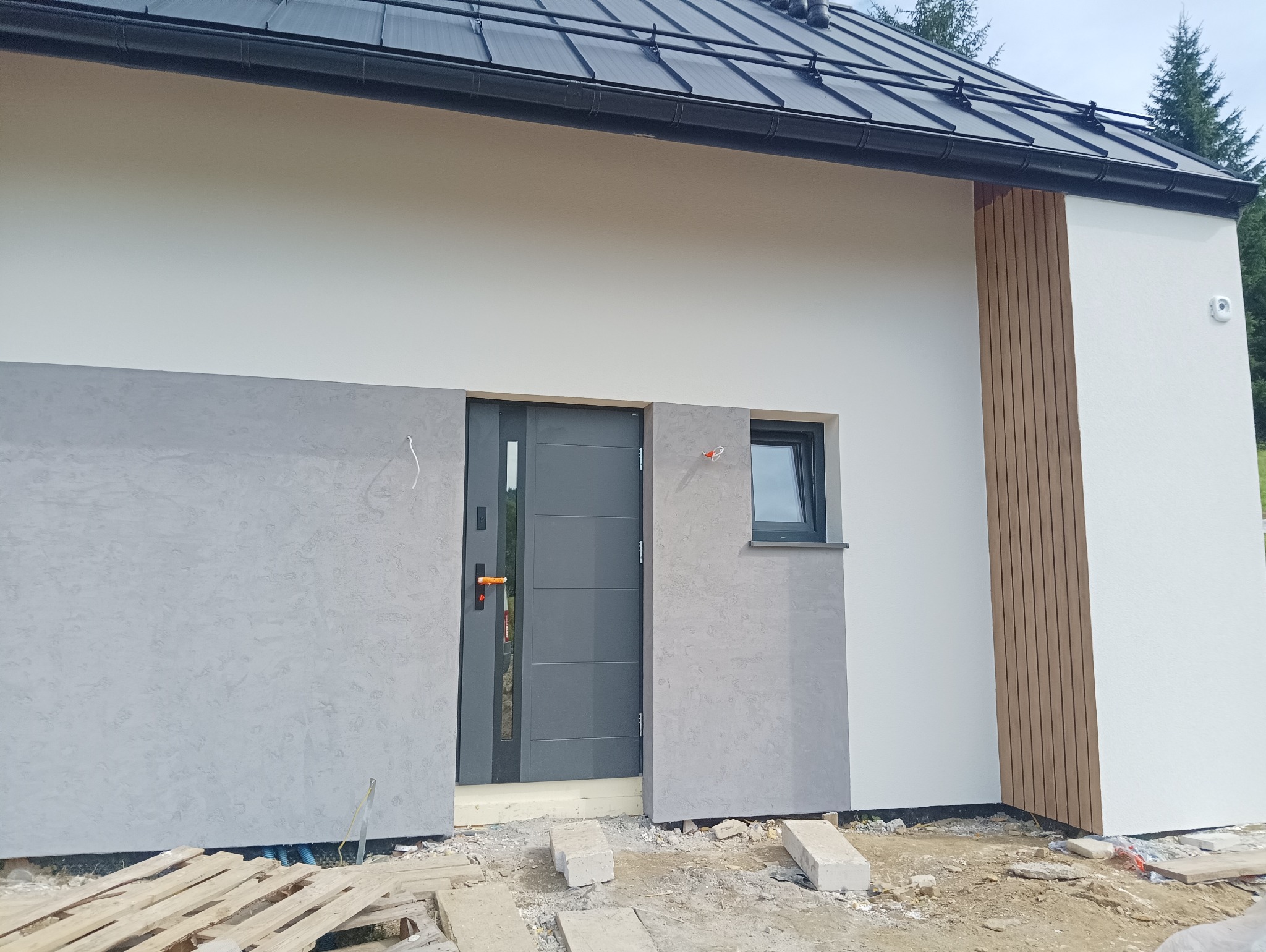 Nowoczesna elewacja domu jednorodzinnego z szarą strukturą tynku wokół drzwi wejściowych i małego okna, uzupełniona drewnianymi panelami pionowymi po prawej stronie, widoczne materiały budowlane...