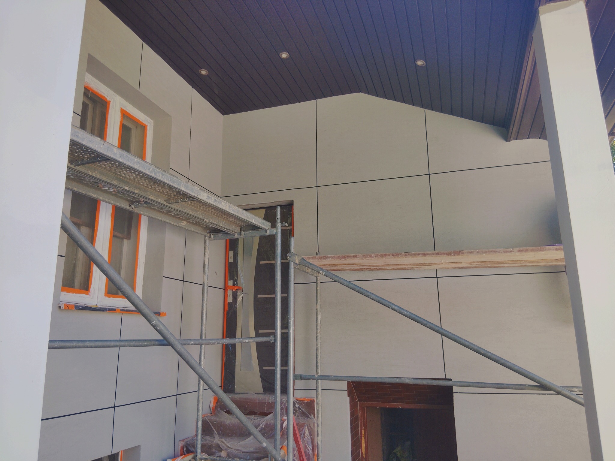 Elewacja budynku w trakcie prac wykończeniowych, widoczne rusztowania, okna zabezpieczone folią, szare panele elewacyjne z ciemnymi fugami, brązowe podbitki.