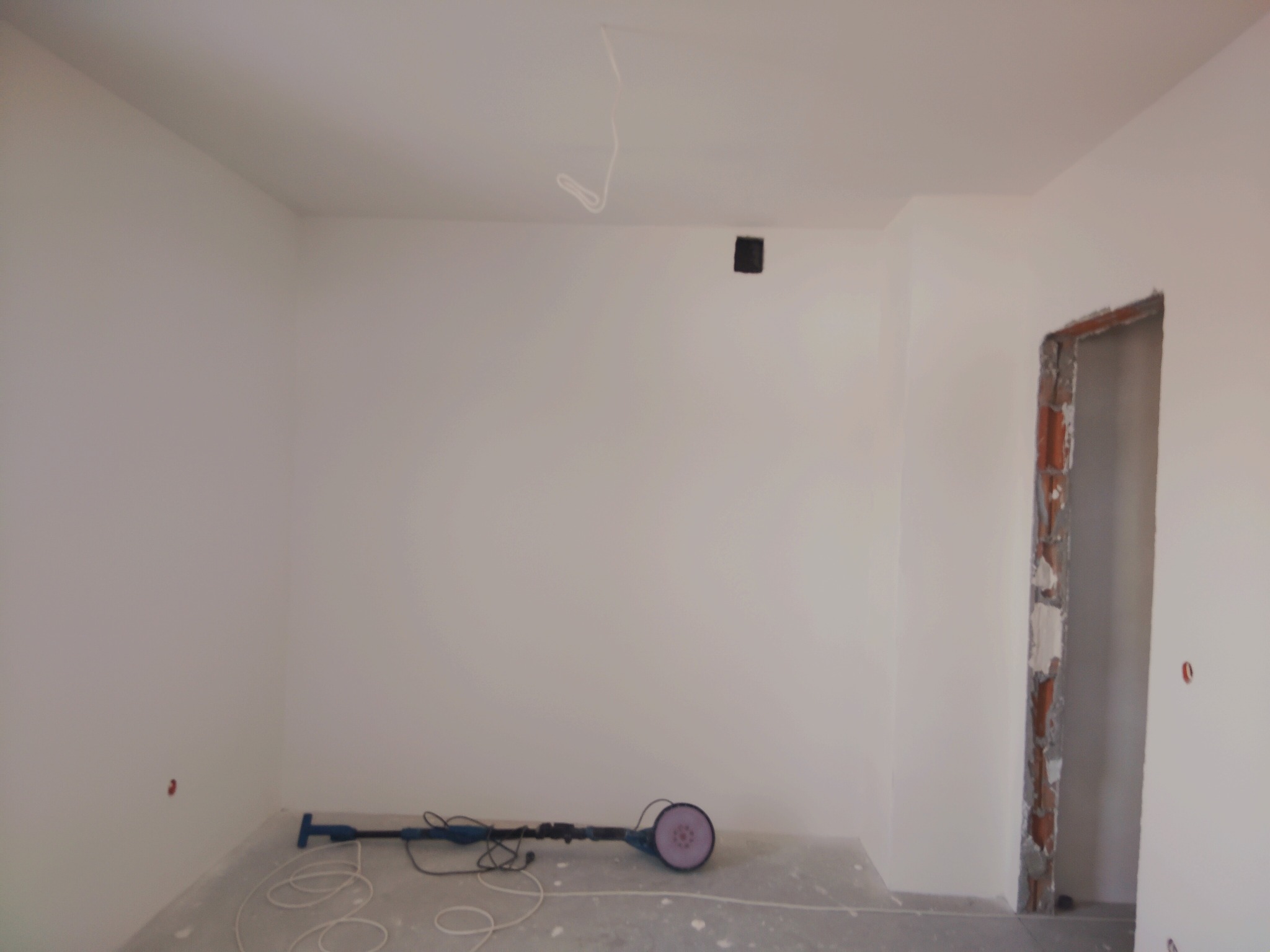 Wnętrze po świeżym malowaniu na biało, z widoczną szlifierką do gładzi na podłodze i surową ościeżnicą drzwiową. Minimalistyczny kadr z detalami wykończeniowymi.