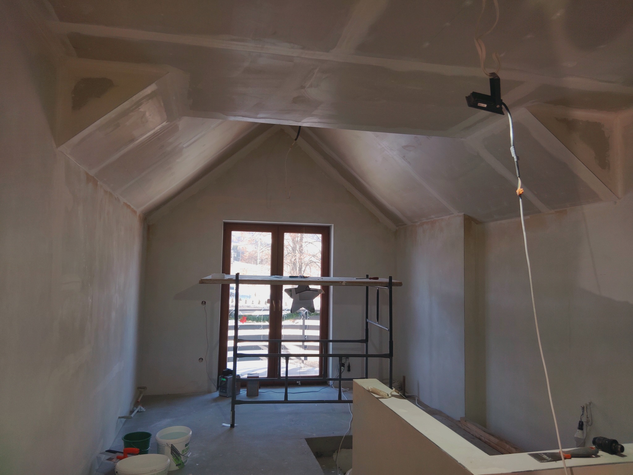 Pokój ze ścianami i sufitem po nałożeniu gładzi, widoczne rusztowanie i narzędzia. Skosy sufitowe i okno w tle. Prace wykończeniowe w jasnym wnętrzu.