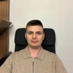 Portret mężczyzny w beżowym swetrze z zamkiem błyskawicznym, siedzącego na czarnym krześle biurowym na tle białej ściany i fragmentu drewnianego regału.